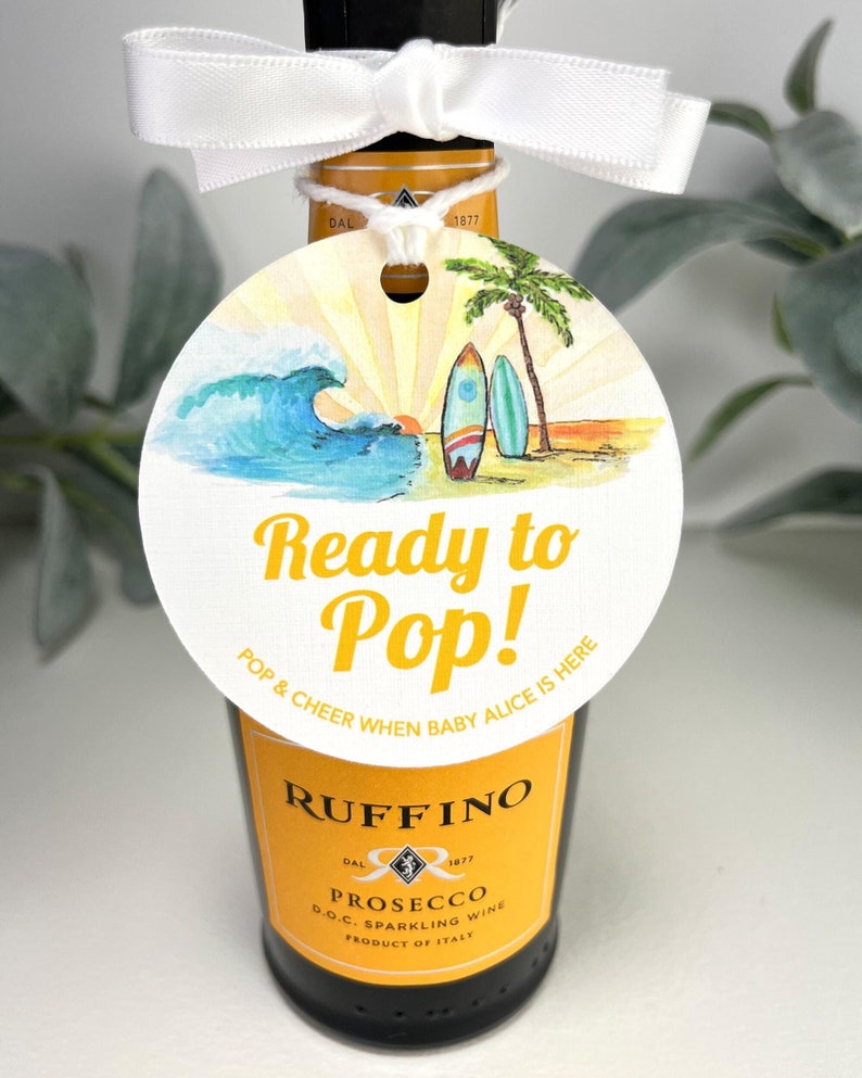 Może przedstawiać: Biała okrągła etykieta z plażową sceną, w tym falą, palmami i deskami surfingowymi. Etykieta zawiera tekst "Ready to Pop!" i "Pop & Cheer When Baby Alice Is Here".