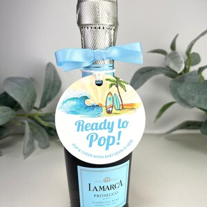 Może przedstawiać: Okrągła etykieta w kolorze niebieskim i białym z plażową sceną i tekstem "Ready to Pop! Pop & Cheer When Baby Kevin Is Here" przymocowana do małej butelki prosecco.