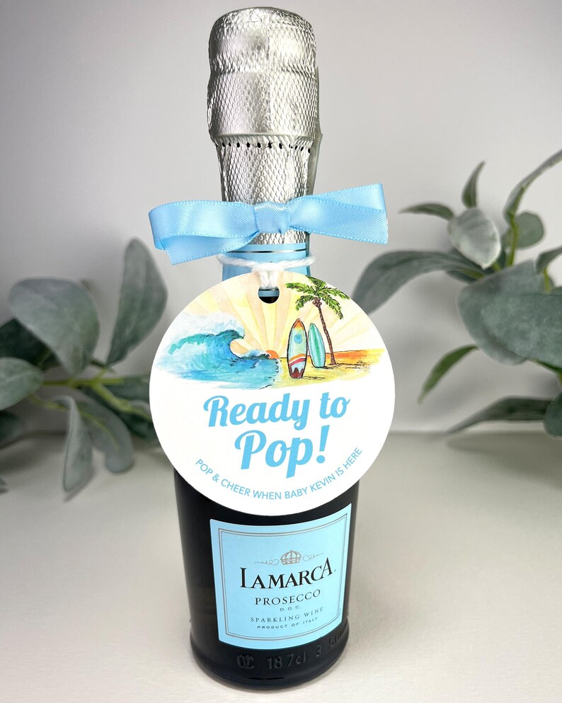 Może przedstawiać: Okrągła etykieta w kolorze niebieskim i białym z plażową sceną i tekstem "Ready to Pop! Pop & Cheer When Baby Kevin Is Here" przymocowana do małej butelki prosecco.