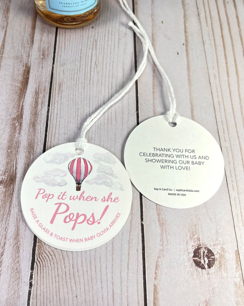 Puede incluir: Dos etiquetas redondas blancas con ilustraciones de globos aerost&aacute;ticos rosas y blancos. El texto en la etiqueta dice "Pop it when she Pops! Raise a glass & toast when baby Olivia arrives". La otra etiqueta dice "Thank you for celebrating with us and showering our baby with love! Say It Card Co. | sayitcardclub.com Made in USA".