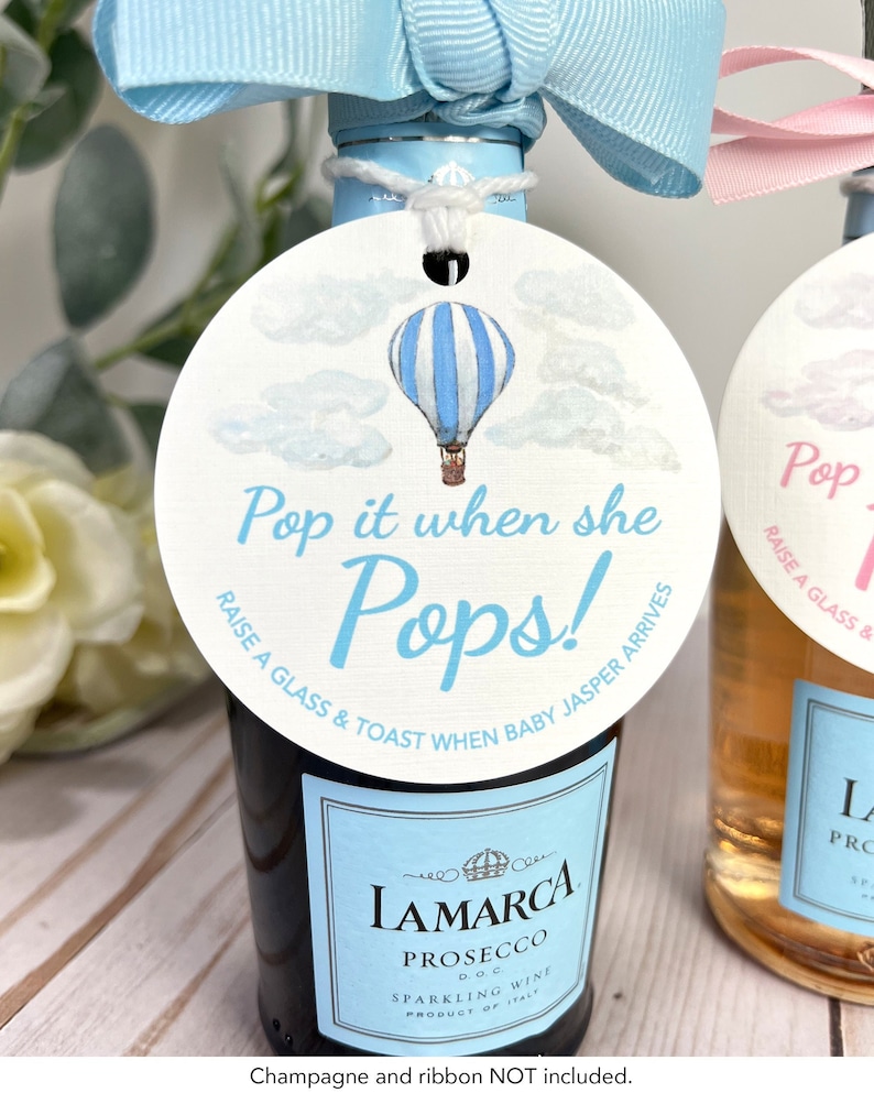 Puede incluir: Una etiqueta circular blanca con una ilustraci&oacute;n de un globo aerost&aacute;tico a rayas azules y blancas. La etiqueta dice "Pop it when she Pops! Raise a glass & toast when baby Jasper arrives."