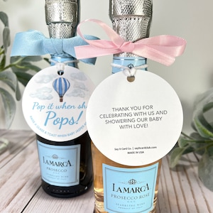 Puede incluir: Dos botellas peque&ntilde;as de vino espumoso con cintas azules y rosas. Las botellas tienen etiquetas adjuntas con el texto "Pop it when she Pops!" y "&iexcl;Gracias por celebrar con nosotros y por dar una fiesta de bienvenida a nuestro beb&eacute; con amor!"