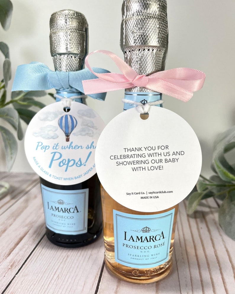 Puede incluir: Dos botellas peque&ntilde;as de vino espumoso con cintas azules y rosas. Las botellas tienen etiquetas adjuntas con el texto "Pop it when she Pops!" y "&iexcl;Gracias por celebrar con nosotros y por dar una fiesta de bienvenida a nuestro beb&eacute; con amor!"