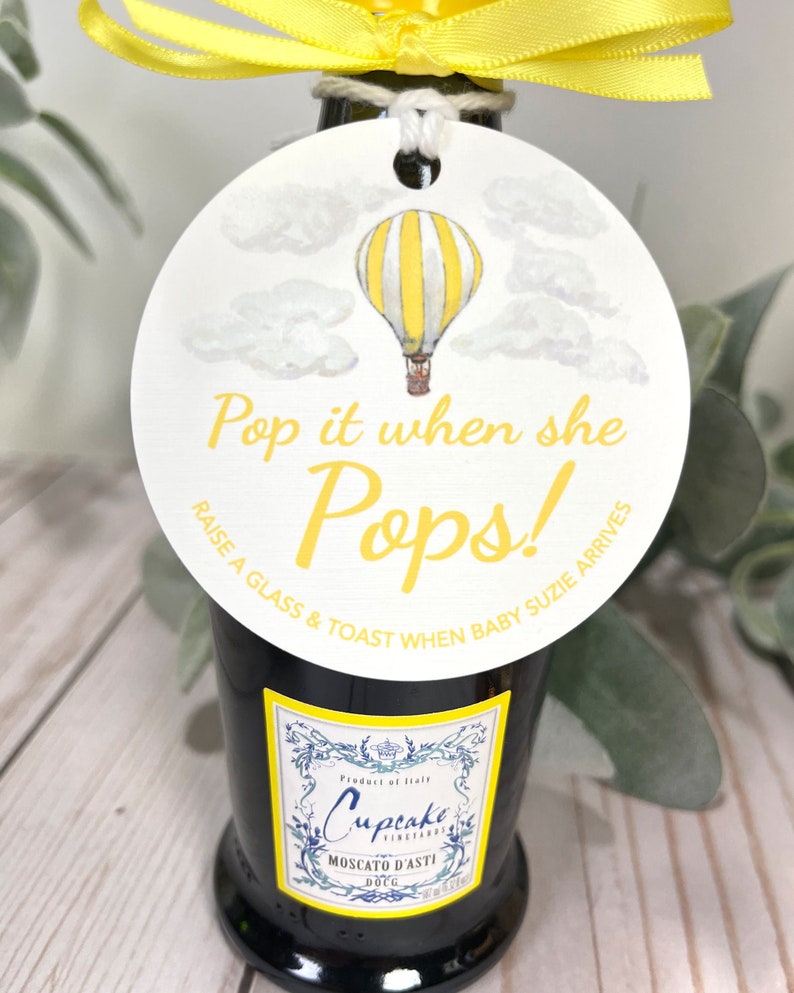 Puede incluir: Una etiqueta redonda blanca con una ilustraci&oacute;n de un globo aerost&aacute;tico a rayas amarillas y blancas. La etiqueta tiene el texto "Pop it when she Pops! Raise a glass & toast when baby Susie arrives."