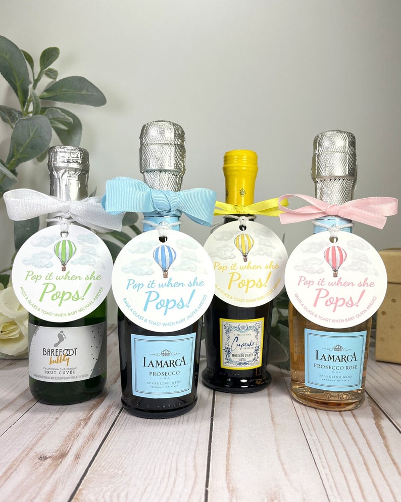 Puede incluir: Cuatro mini botellas de vino espumoso con etiquetas personalizadas que dicen "Pop it when she Pops!"  Brinda con una copa y celebra la llegada del beb&eacute;. Las etiquetas presentan un dise&ntilde;o de globo aerost&aacute;tico con detalles en azul, amarillo y rosa.