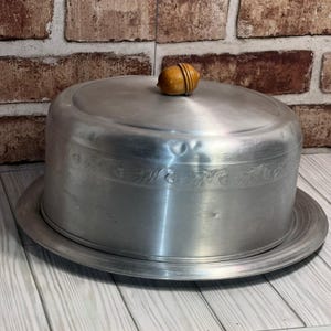 Könnte beinhalten: Eine runde Tortenhaube aus gebürstetem Aluminium mit passendem Tablett. Die Haube ist mit einem horizontalen Band mit geprägtem Blatt- und Eichelmotiv verziert. Ein kleiner, glatter, hellbrauner Holzknauf in Eichelform sitzt oben auf dem Deckel.