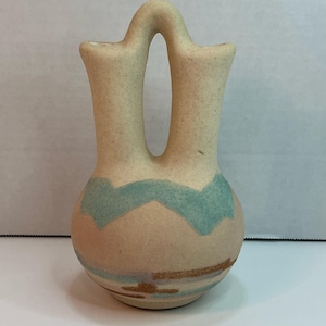 May include: A tan ceramic vase with a distinctive double-spout design. The vase has a turquoise zigzag pattern around the middle and brown horizontal lines near the base. The vase has a smooth, matte finish and stands upright.