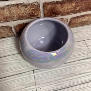 May include: A spherical ceramic bowl with a smooth, light lavender finish and a subtle iridescent sheen that reflects soft rainbow colors. The interior is hollow and finished in a matching light lavender glaze, featuring a wide, circular opening.