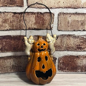 May include: A glossy orange ceramic jack-o'-lantern lantern featuring a carved face with triangular eyes and a wide grin. Two small, light-colored ghost figures are attached to the sides, and a thin black metal wire handle is attached to the top.