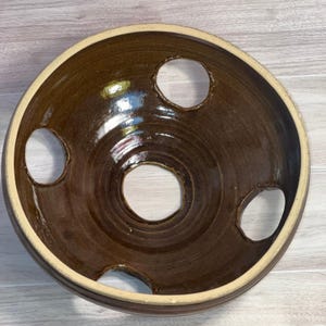 May include: A round, dark brown glazed ceramic bowl featuring a smooth, glossy finish and a light-colored rim. The bowl is designed with four circular openings: one large hole centered at the base and three smaller holes spaced along the sides.
