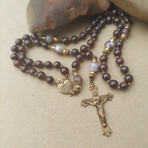 Può includere: Un rosario con perline rosso scuro e dettagli color oro. Il rosario presenta un crocifisso e un ciondolo a forma di cuore. Le perline sono di un colore rosso intenso e i componenti metallici hanno un aspetto vintage.