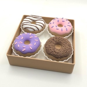 May include: A box of four decorative donuts. One donut has white icing with brown stripes, another has pink icing with purple sprinkles, a third has purple icing with pink sprinkles, and the last has brown chocolate frosting.