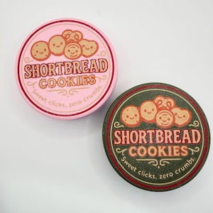 May include: Two round containers of shortbread cookies. One is pink with red accents, the other is green with red accents. Both feature the text "SHORTBREAD COOKIES" and the phrase "Sweet clicks, zero crumbs."