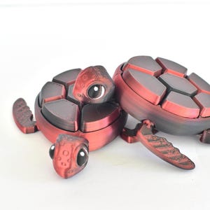 May include: Two red and black 3D-printed turtle figurines. The turtles have segmented shells, detailed flippers, and large, expressive eyes. The figurines are displayed on a white background.