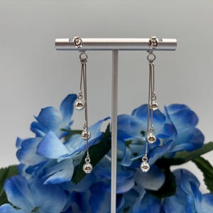 May include: Silver dangle earrings with three small, round, silver beads suspended from a thin silver bar. The earrings are displayed on a silver stand against a backdrop of blue hydrangea flowers.