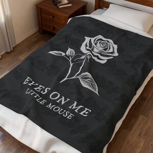May include: A black and white floral blanket with a large rose illustration and the text "EYES ON ME LITTLE MOUSE". The blanket is on a bed with a white pillow and a wooden nightstand.