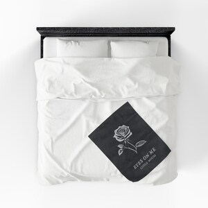 May include: A black throw blanket with a white rose illustration and the text "EYES ON ME LITTLE MOUSE" is displayed on a white bed with white bedding. The bed frame is black.