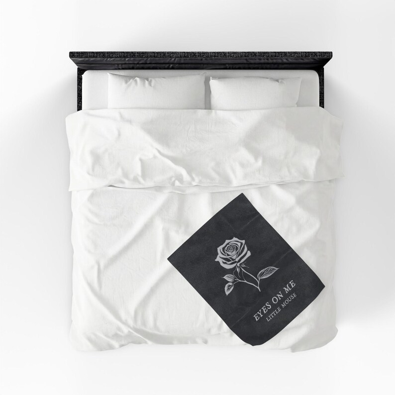 May include: A black throw blanket with a white rose illustration and the text "EYES ON ME LITTLE MOUSE" is displayed on a white bed with white bedding. The bed frame is black.