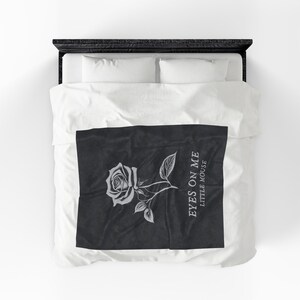 May include: A black and white blanket featuring a detailed rose illustration with leaves and the text "EYES ON ME LITTLE MOUSE". The design is centered on the blanket, which is placed on a bed with white bedding.