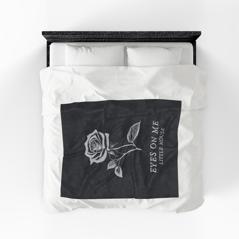 May include: A black and white blanket featuring a detailed rose illustration with leaves and the text "EYES ON ME LITTLE MOUSE". The design is centered on the blanket, which is placed on a bed with white bedding.