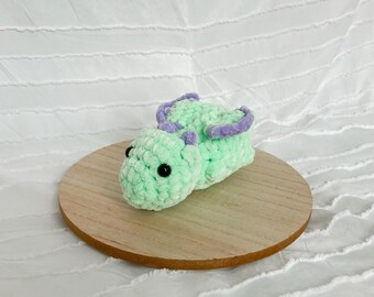 Puede incluir: Un pequeño dragón verde de crochet con alas moradas. El dragón está acostado de lado con la cabeza apoyada en sus patas delanteras. El dragón tiene ojos negros y una expresión linda.
