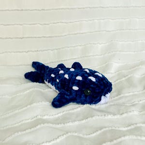 May include: A handmade plush whale shark in dark blue with white spots. The stuffed animal has a white belly and a black button eye. The plush toy is lying on a white, textured surface.