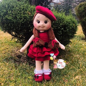 Könnte beinhalten: Eine gehäkelte Puppe, die ein rotes Kleid, eine rote Baskenmütze und rote Schuhe trägt. Die Puppe hat braunes Haar in Zöpfen und ein lächelndes Gesicht.