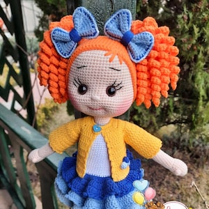Könnte beinhalten: Eine gehäkelte Puppe mit orangefarbenen Locken, blauen Schleifen, einem gelben Cardigan, einem blau-weißen Kleid und blauen Schuhen. Die Puppe steht auf einem Holzzaun.