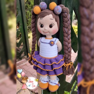Könnte beinhalten: Eine gehäkelte Puppe mit braunen Haaren, einem weißen Kleid mit einer lila Blume und einem lila-orangenen Rock. Die Puppe steht neben einem Holzzaun.