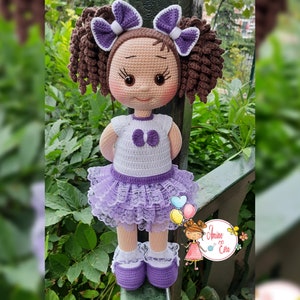 Könnte beinhalten: Eine gehäkelte Puppe mit braunen Locken, einem lila-weißen Kleid und lila Schuhen. Die Puppe trägt eine lila Schleife mit weißem Zentrum.