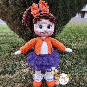 Könnte beinhalten: Eine gehäkelte Puppe mit braunen Haaren, die einen orangefarbenen Cardigan, ein weißes Hemd, einen lila Rock mit Spitzenbesatz und orangefarbene Schuhe trägt. Die Puppe steht auf grünem Gras.