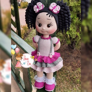 Könnte beinhalten: Eine gehäkelte Puppe mit schwarzen Locken, die ein rosa-graues Kleid mit einem weißen Häkelkragen und ein Paar rosa Schuhe trägt.