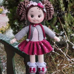 Könnte beinhalten: Eine gehäkelte Puppe mit braunen Haaren, einem rosa-weißen Blumen-Haarband, einem grauen Cardigan, einem rosa Kleid und weißen Schuhen. Die Puppe steht auf einem Holzgeländer.
