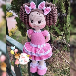 Könnte beinhalten: Eine gehäkelte Puppe mit braunen Haaren, einem rosa Kleid und weißen Schuhen. Die Puppe steht vor einem Holzzaun.
