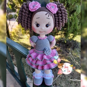 Könnte beinhalten: Eine gehäkelte Puppe mit braunen Locken, rosa Blumen, einem grauen Kleid und einem rosa Rüschenrock. Die Puppe trägt graue Schuhe und weiße Socken.