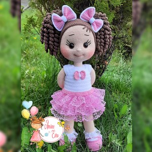 Könnte beinhalten: Eine gehäkelte Puppe mit braunen Haaren, rosa Schleifen und einem rosa-weißen Kleid. Die Puppe steht auf grünem Gras.