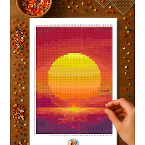 May include: A colorful pixel art project depicting a sunset with a large yellow sun and a purple and red sky. The project is on a white sheet with a grid, surrounded by colorful beads and tools on a wooden surface.