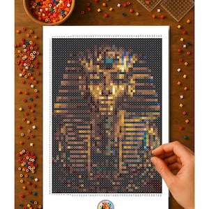 May include: A detailed pixel art image of the bust of Tutankhamun, rendered in shades of brown, gold, and blue. The artwork is on a white sheet, with a hand placing a bead. A bowl of colorful beads and pegboards are on a wooden surface.