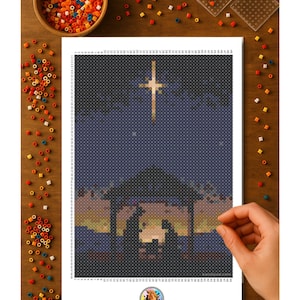 May include: A completed bead art project depicting a nativity scene with a cross in the night sky. The artwork is on a white sheet, surrounded by colorful beads and tools on a wooden surface. The Two Suns logo is visible.