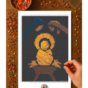 May include: A pixel art design of a nativity scene, featuring a baby in a manger, with hands reaching down from above. The image is on a white sheet, surrounded by colorful beads and crafting tools, all on a wooden surface.