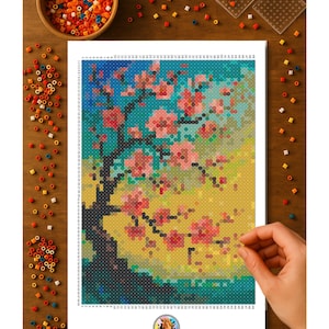 May include: A colorful pixel art of a cherry blossom tree with pink flowers and a dark trunk. The artwork is on a white sheet, with a hand placing a bead. A wooden bowl and scattered beads are on the wooden surface.