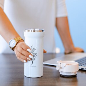 May include: A white stainless steel travel mug with a black floral design. The mug is sitting on a wooden table next to a laptop computer.