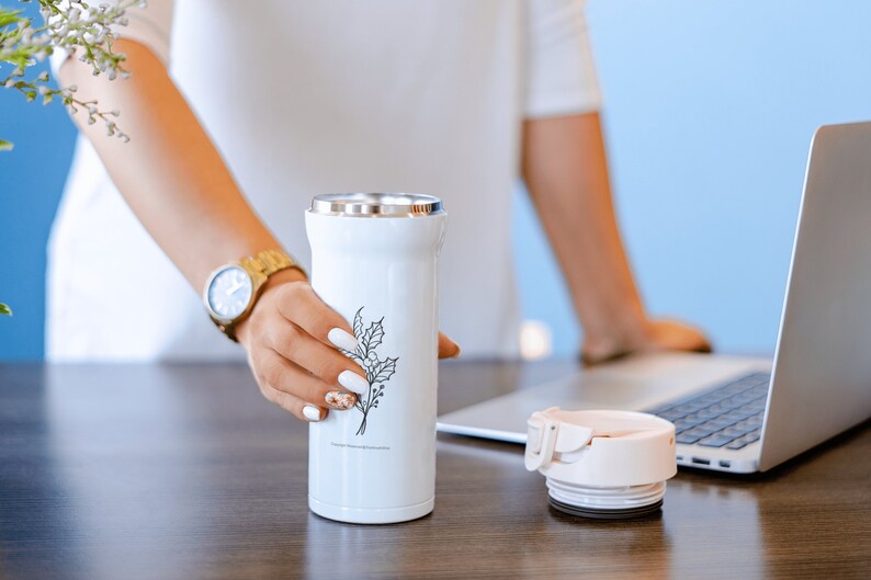 May include: A white stainless steel travel mug with a black floral design. The mug is sitting on a wooden table next to a laptop computer.