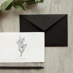 May include: A white card with a black line drawing of a holly sprig on a marble surface with a black envelope and a black pencil.