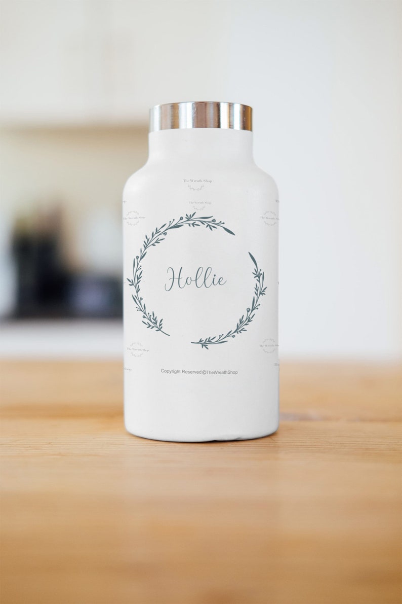 May include: White insulated water bottle with a silver lid and a green floral wreath design. The name "Hollie" is written in the centre of the wreath. The text "Copyright Reserved @TheWreathShop" is printed at the bottom of the bottle.