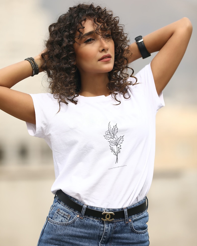 May include: A white t-shirt featuring a black floral design and a black belt with a gold buckle. The model is wearing blue jeans.