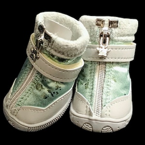 May include: A pair of light green dog boots with white soles and silver zippers. The boots have a white strap with a silver paw print charm.