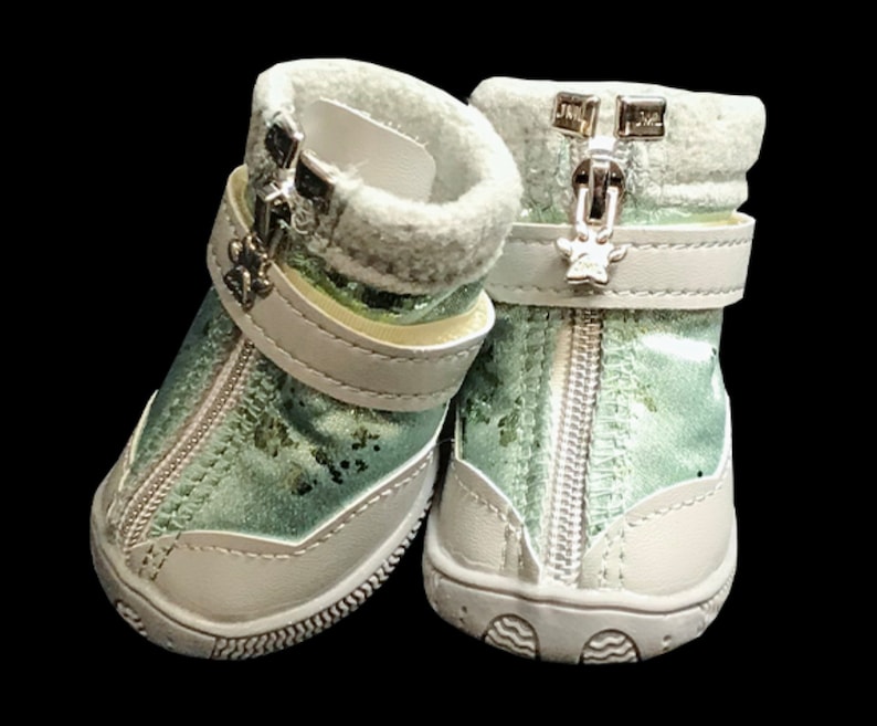 May include: A pair of light green dog boots with white soles and silver zippers. The boots have a white strap with a silver paw print charm.
