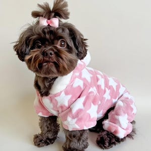 May include: A small, dark-haired dog wearing a pink and white star-patterned onesie with a hood. The dog has a pink bow in its hair. The onesie covers the dog's legs and torso, providing warmth and style.