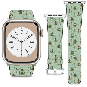 May include: A light green Apple Watch strap featuring a repeating pattern of cartoon frogs, lily pads, and reeds. The watch face displays the time. The strap is shown separately and attached to the watch.